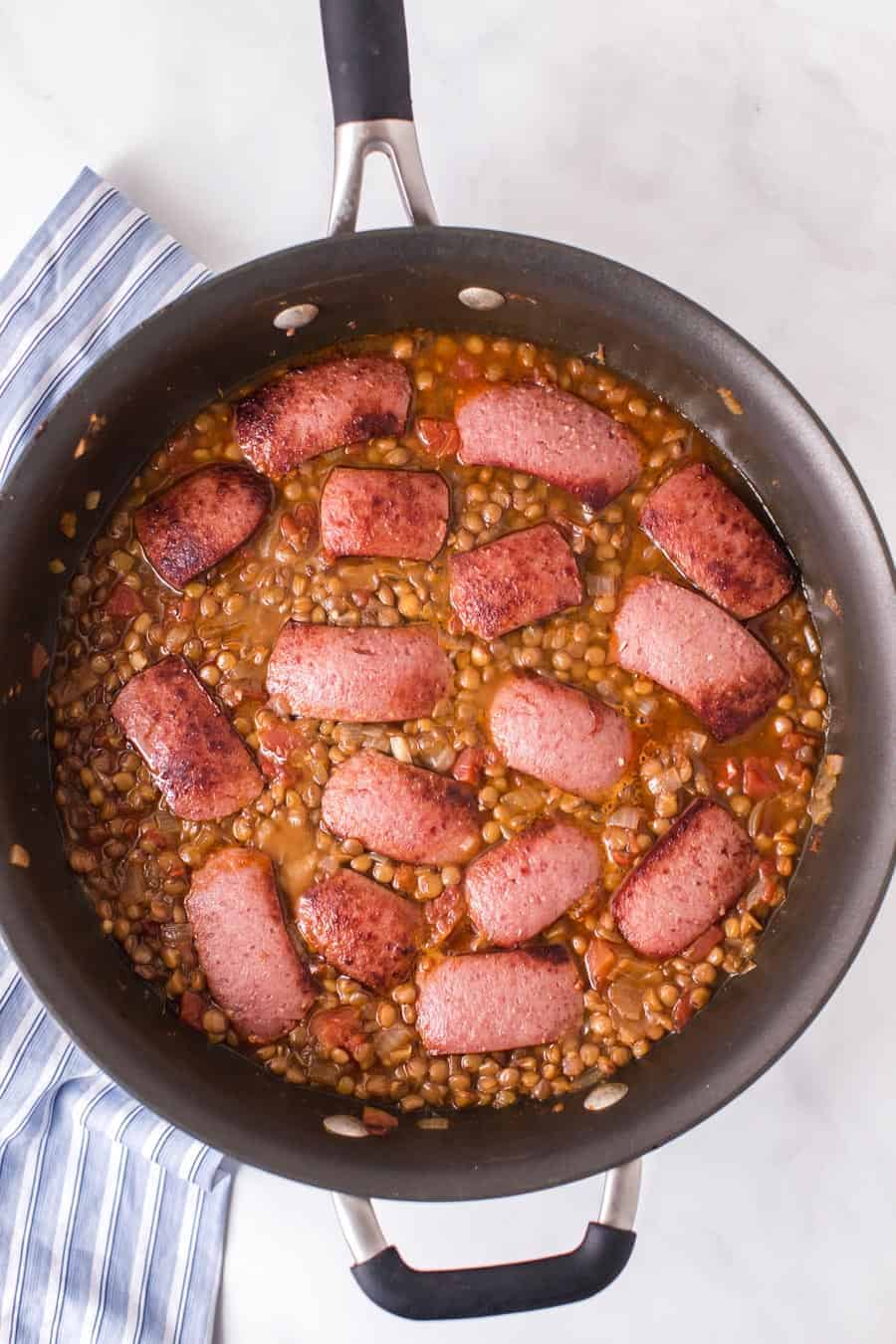 lentils cooking with link sausage that has been browned floating on top in a pan.