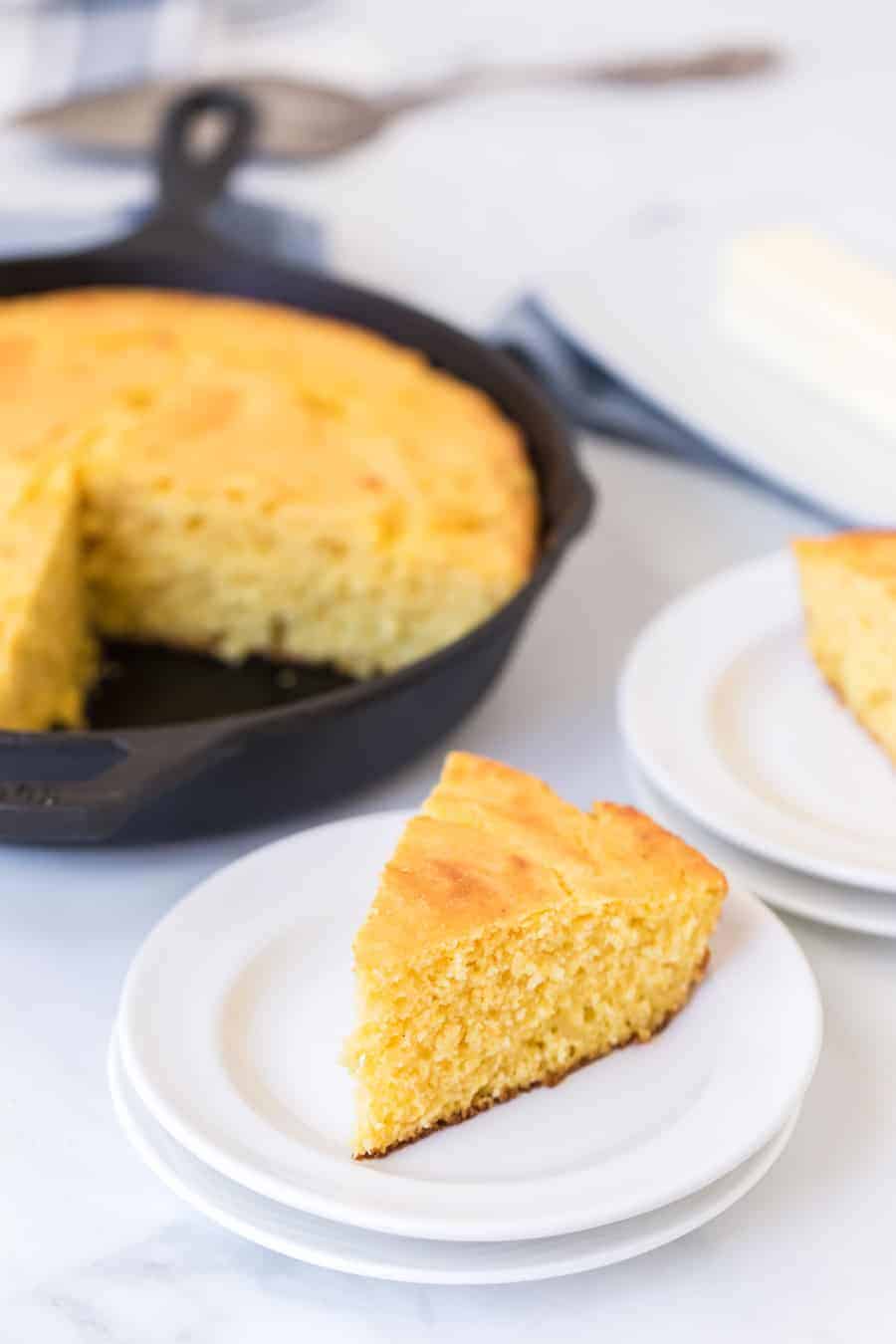 cornbread baked in a cast iron skillet in the background then triangle pieces have been served on white plates.