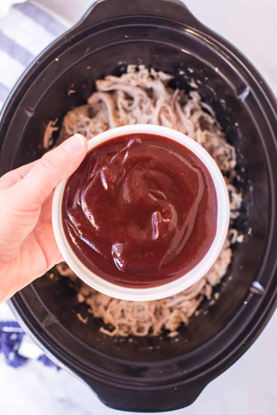 pork shredded in an instant pot and hand holding a white dish of BBQ sauce over it, ready to pout in.