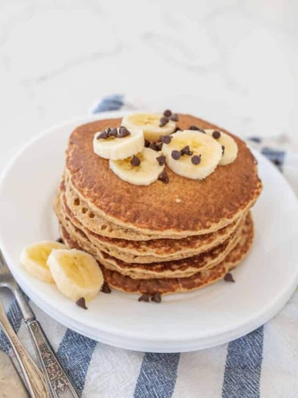 stack of banana pancakes on round white plate with banana slices on top of blue striped towel