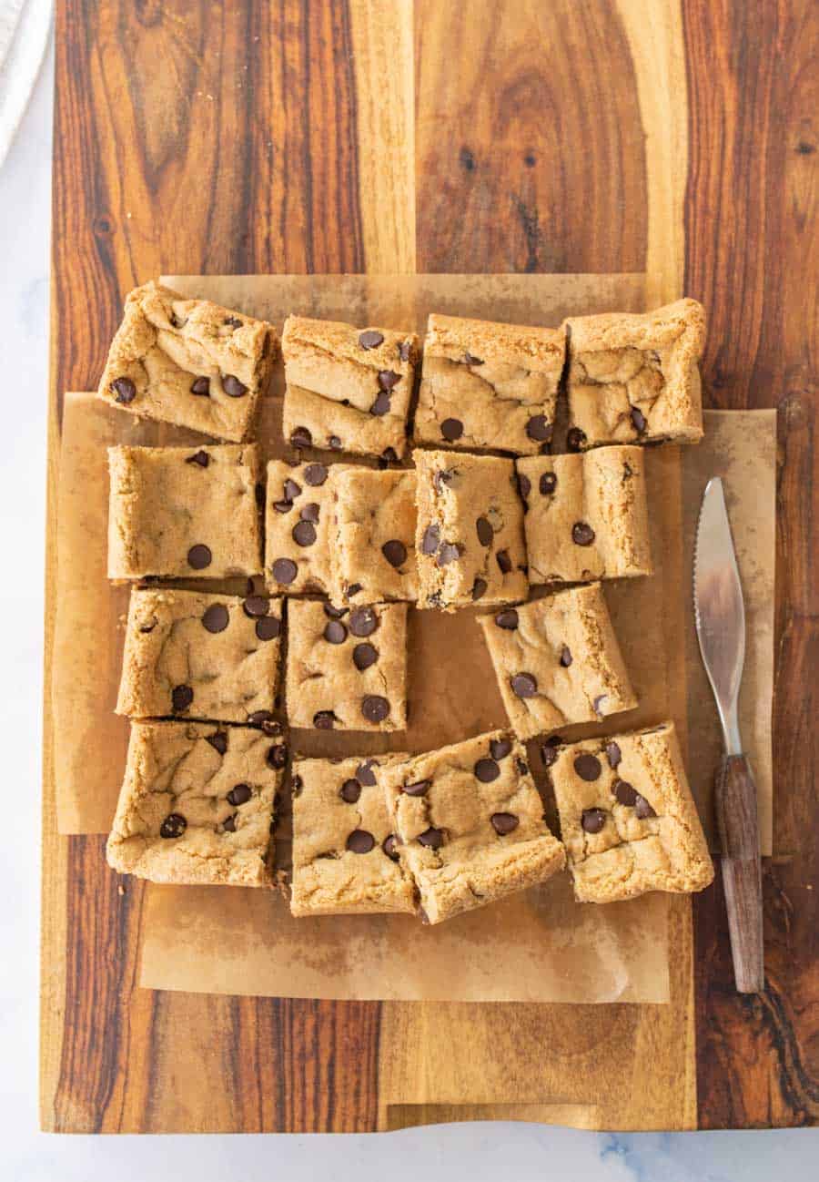 Baked cookie bars cut into squares on top of parchment paper on a wooden cutting board with a knife next to them. 