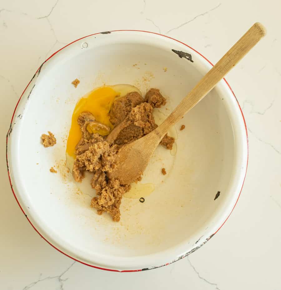 Egg and vanilla added to the butter and sugar mixture with a wooden spoon in a white bowl with a red rim on a white marble counter. 