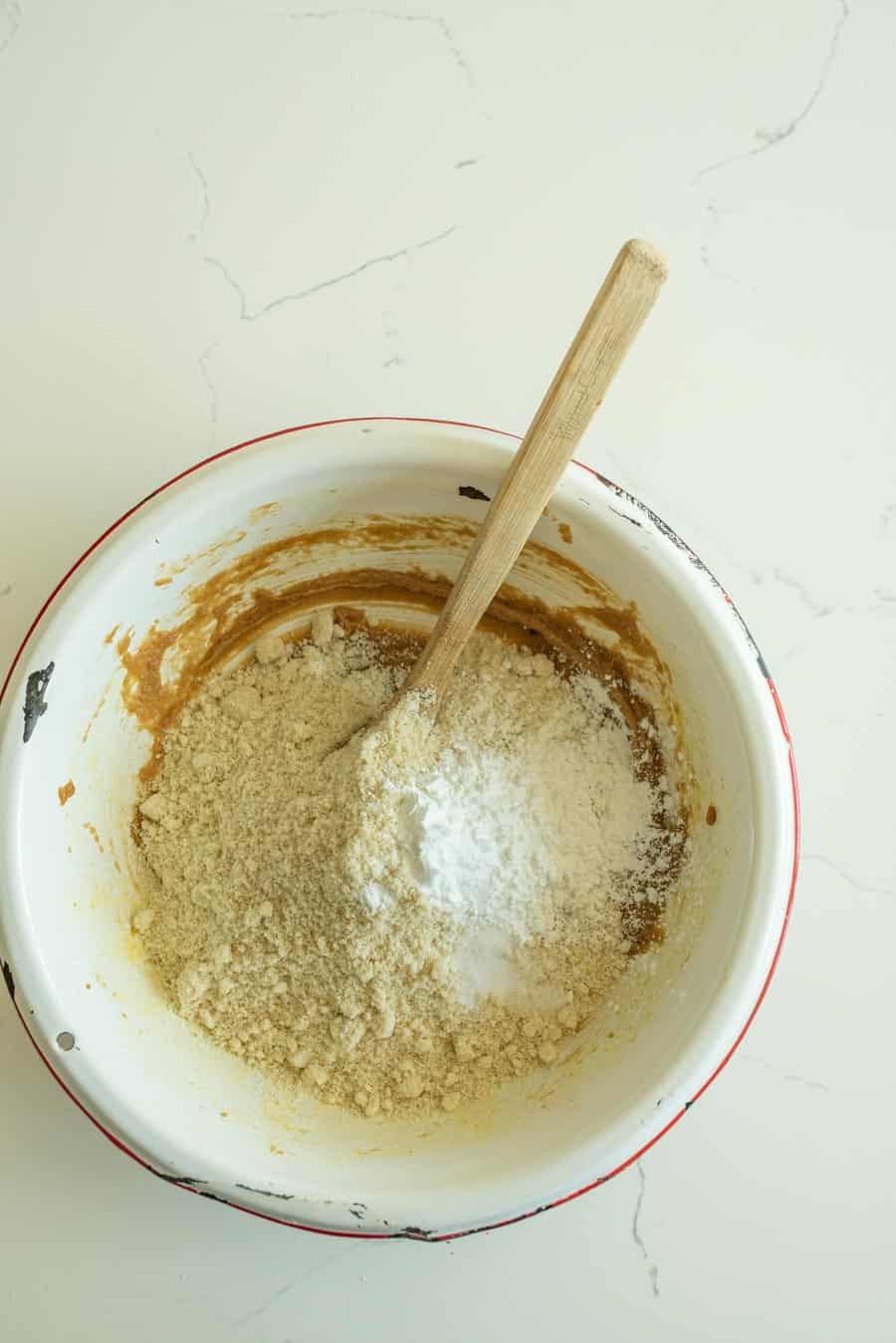 Almond flour and dry ingredients added to the cookie bar dough with a wooden spoon in a white bowl with a red rim. 