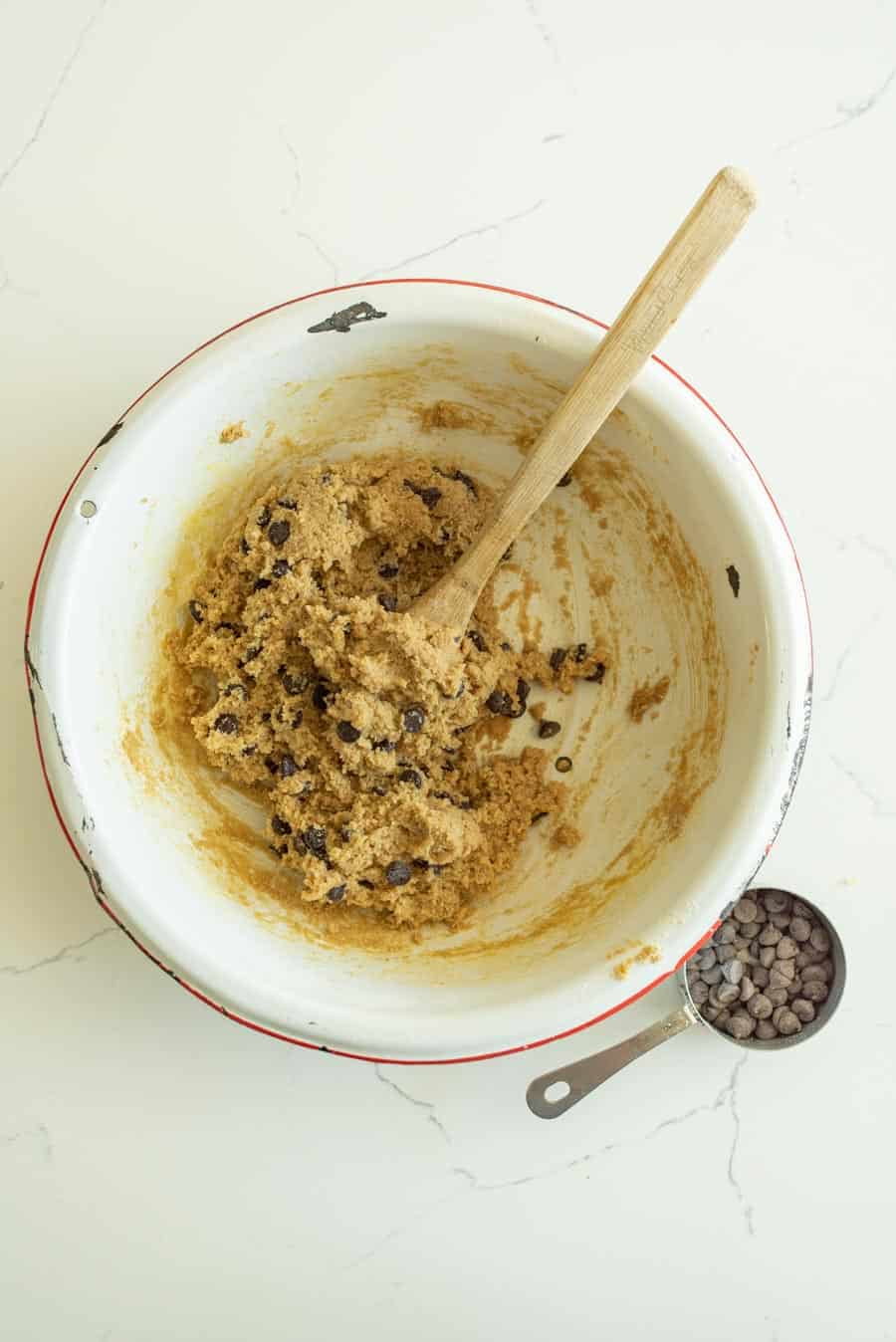 Almond flour cookie bar dough with the chocolate chips mixed in with a wooden spoon in a white bowl with a red rim on a white marble counter. 