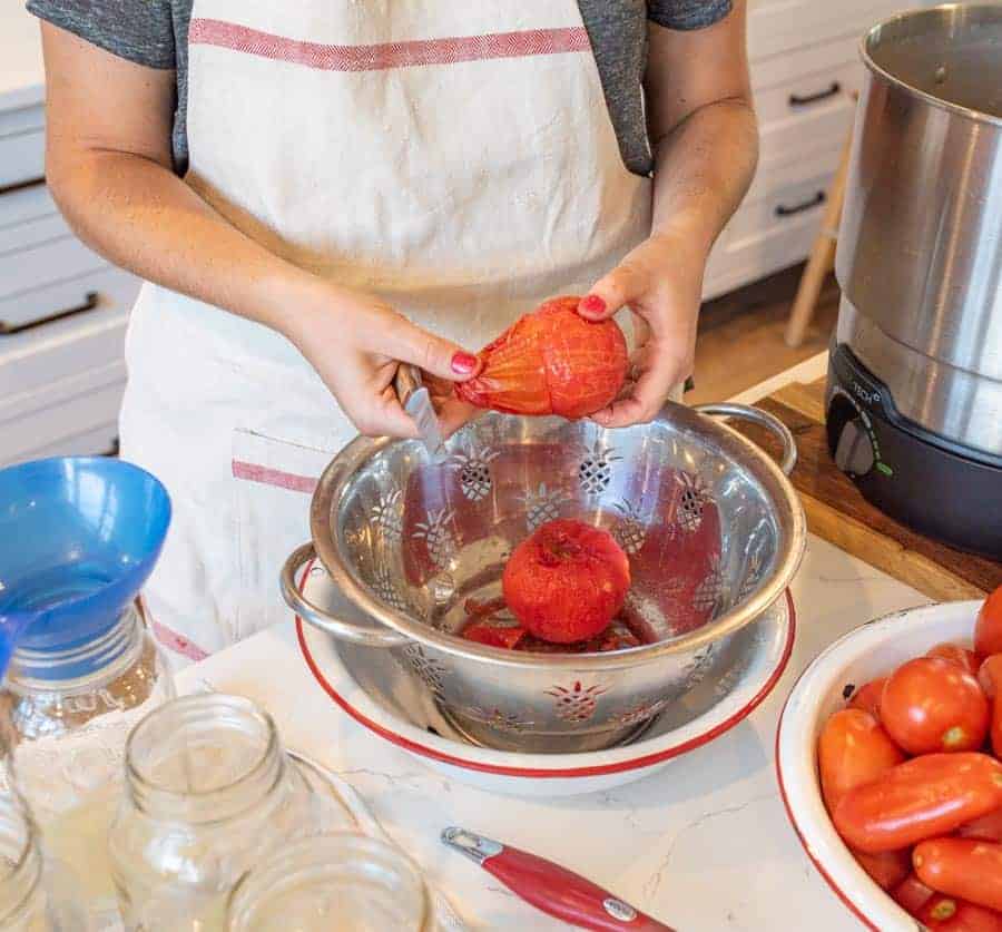 A woman peeling the skin off of a blanched tomatoes above a colander. 