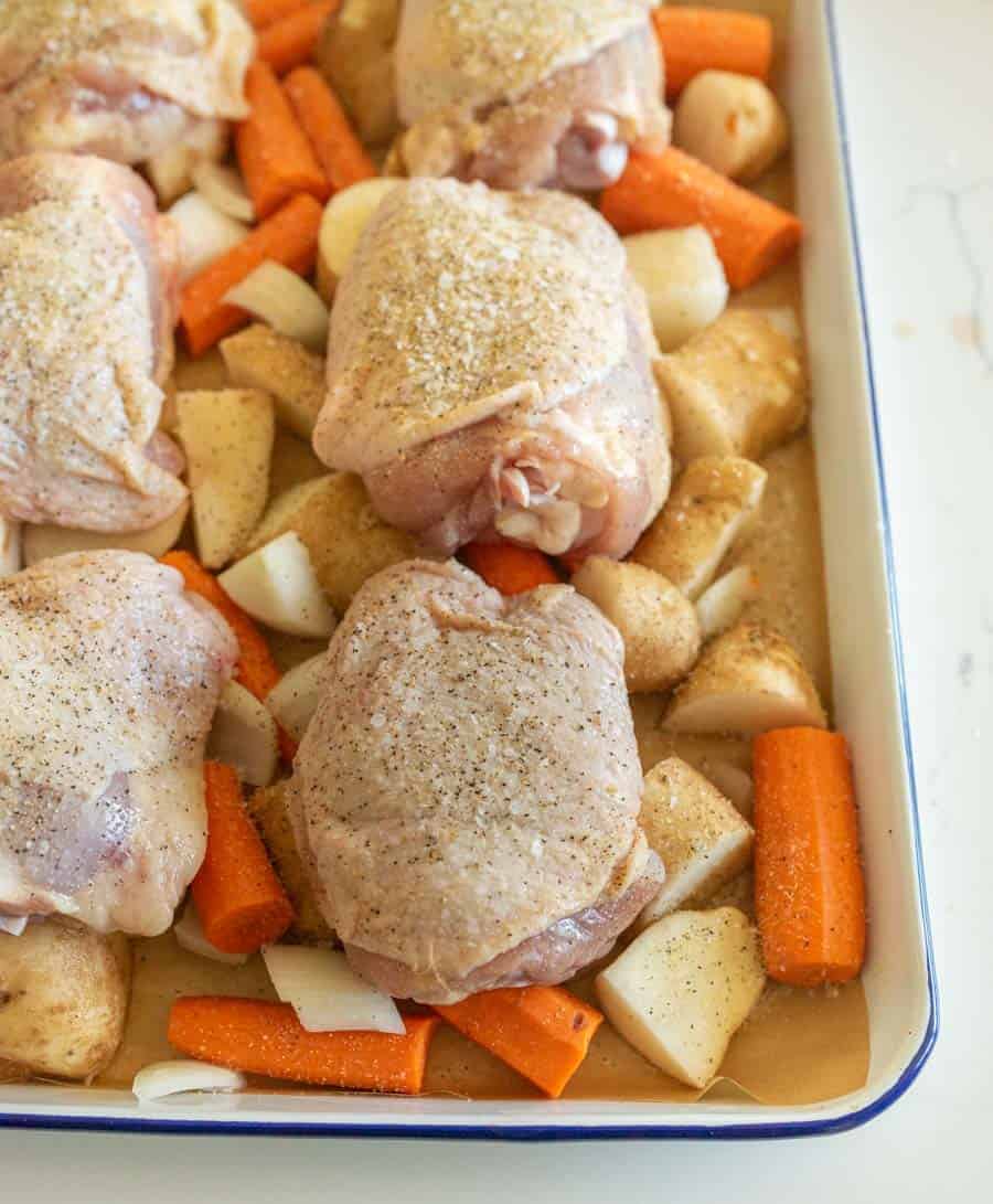 Close-up of chicken thighs, chopped carrots, potatoes, and onions on a lined enamel baking sheet ready for the oven.