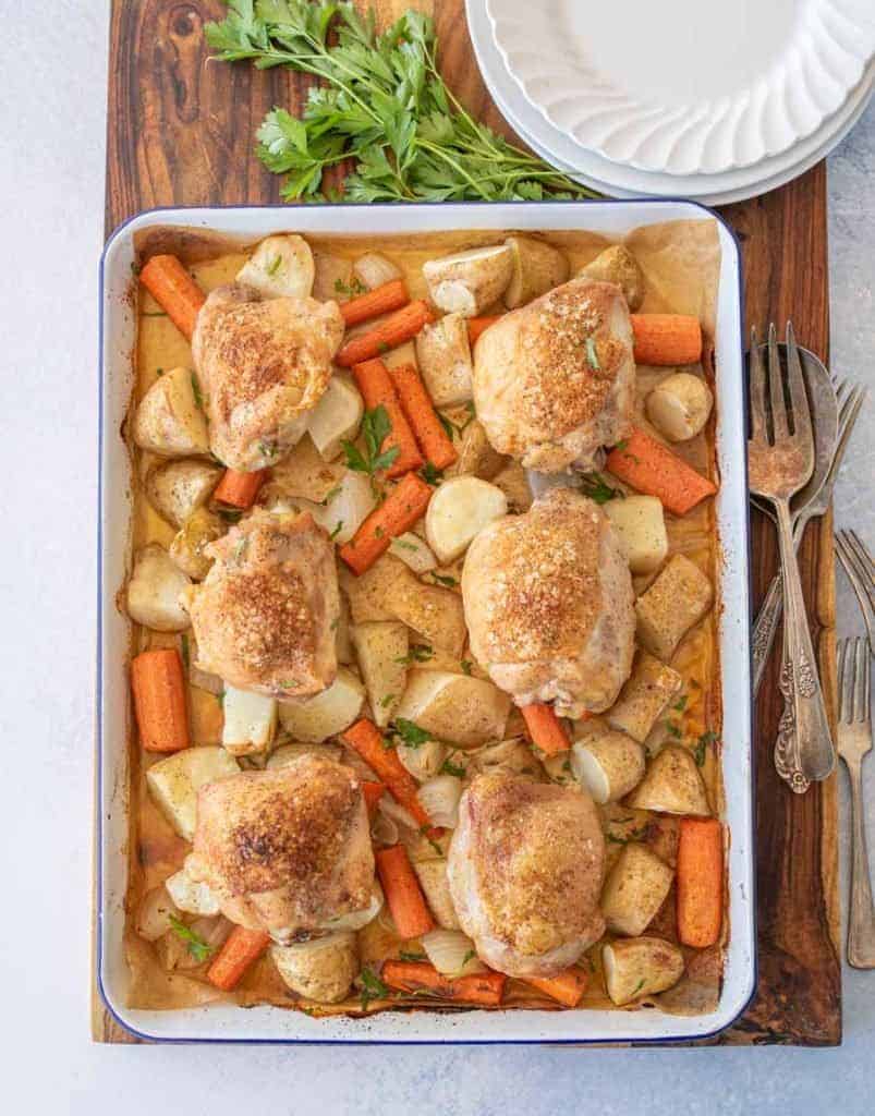 oven roasted chicken thighs, carrots, potatoes, and onions on enamel baking sheet with dinner plates, forks, and garnish