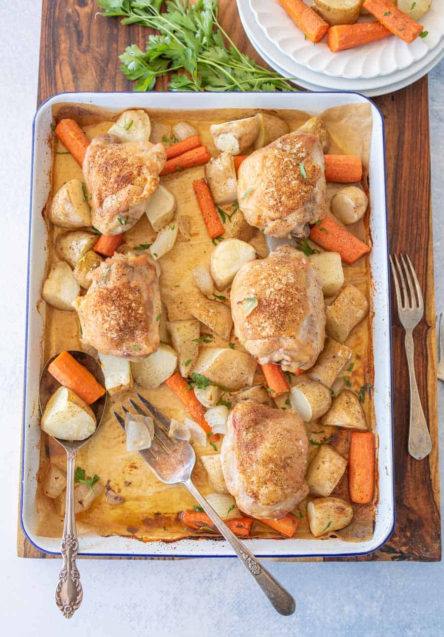 Five oven roasted chicken thighs, carrots, potatoes, and onions on a lined enamel baking sheet after coming out of the oven next to a dinner plate and fork.