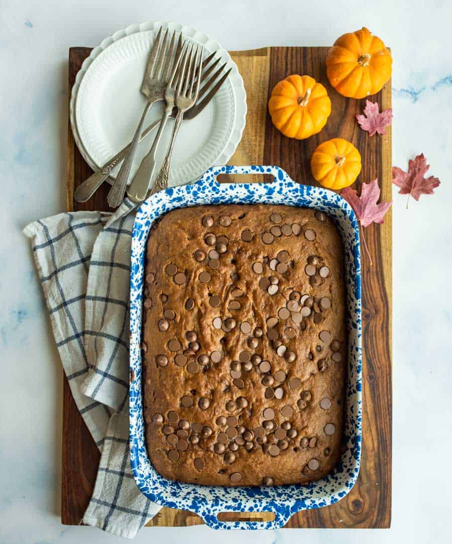 A whole baked chocolate chip pumpkin cake in a blue pan with two white plates, four forks and three pumpkins next to the pan on a wooden board. 