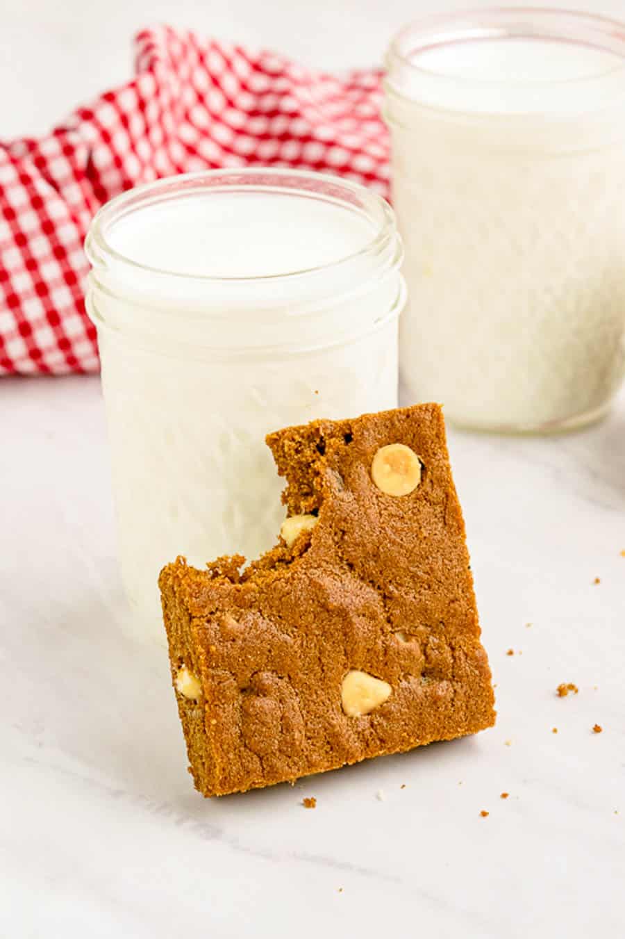 A gingerbread blondie bar with a bite taken out it leaning against a glass of milk.