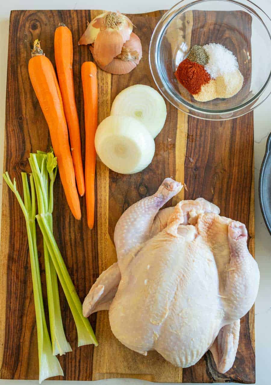 All of the ingredients to roast a whole chicken on a wooden cutting board including carrots, celery, onions, spices, and a whole raw chicken. 