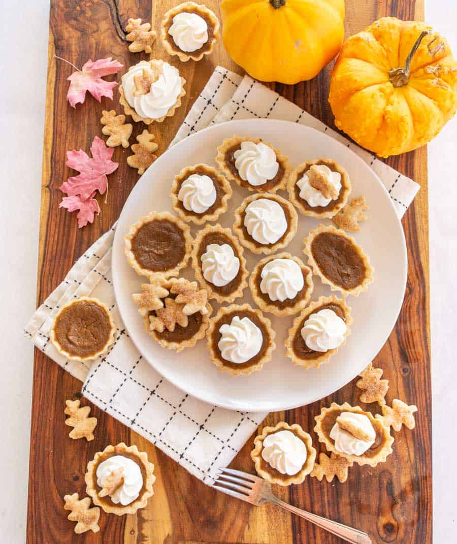 Mini pumpkin pies with whipped cream swirl on top on a white plate on a wooden cutting board next to two pumpkins.