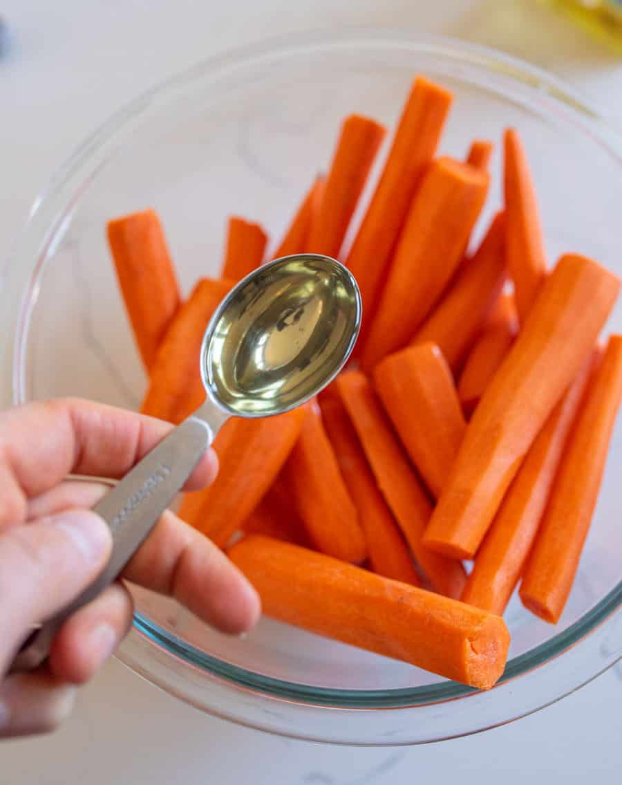 Cut carrots in a clear glass bowl with a hand holding a measuring spoon with oil. 