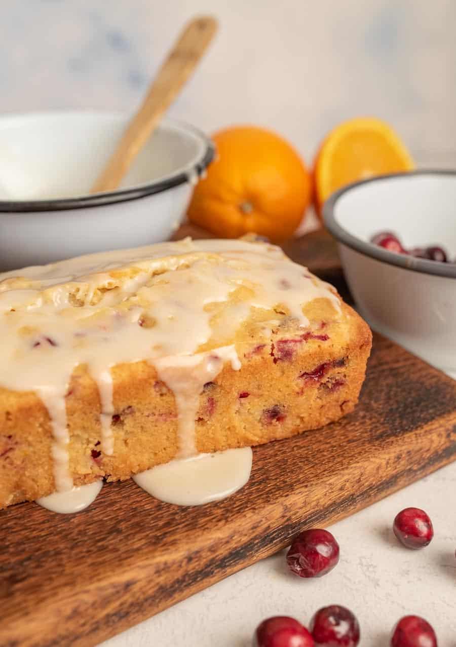 Close-up of the side of a loaf of cranberry orange bread with the glaze dripping down the side with a couple oranges, a bowl of cranberries, and the bowl of glaze in the background. 