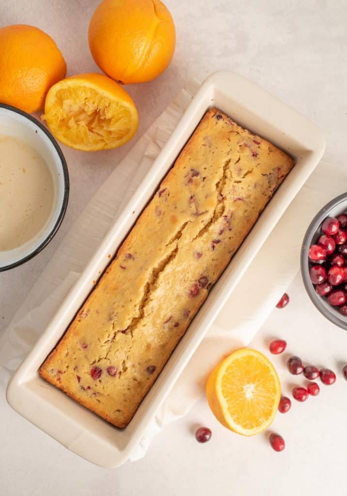 loaf of cranberry orange bread in white loaf pan surrounded by oranges and cranberries