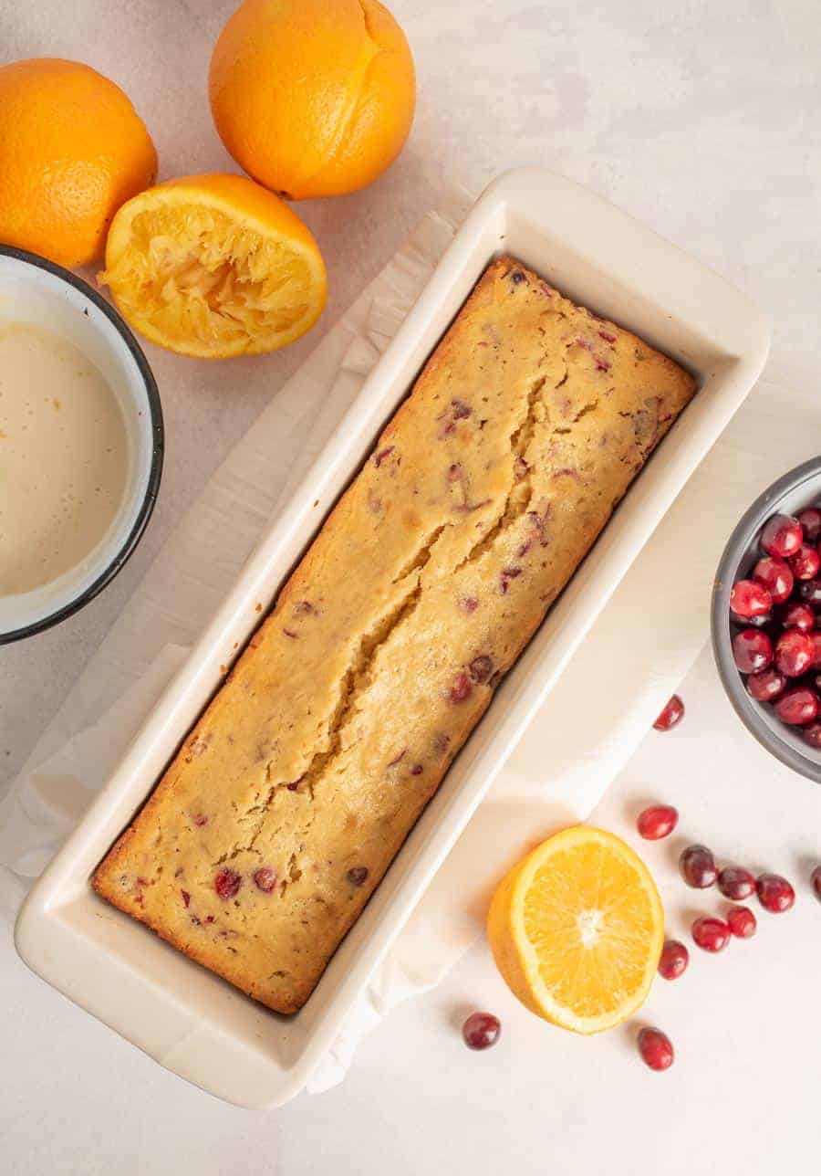 Baked cranberry orange bread in a white ceramic tea cake pan on a white counter next to an orange cut in half and a bowl of cranberries.