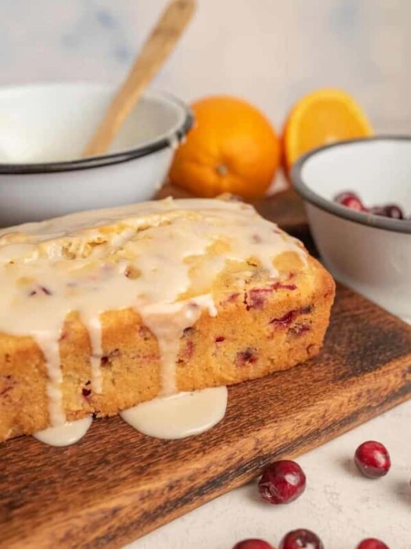 loaf of cranberry orange bread with orange glaze drizzled on top surrounded by cranberries, bowls, and oranges