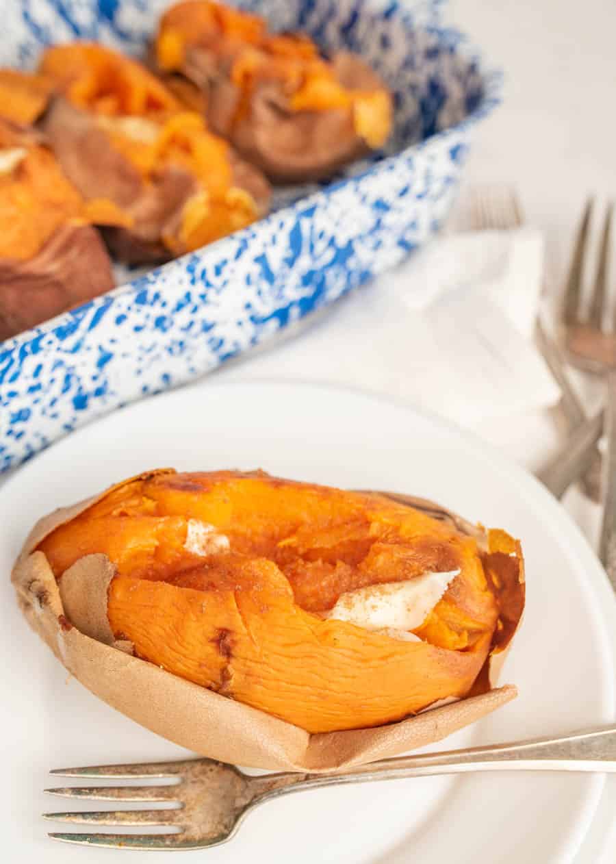 A baked sweet potato with butter on it on a white plate with a fork and three baked sweet potatoes in the background in a white and blue speckled baking dish.