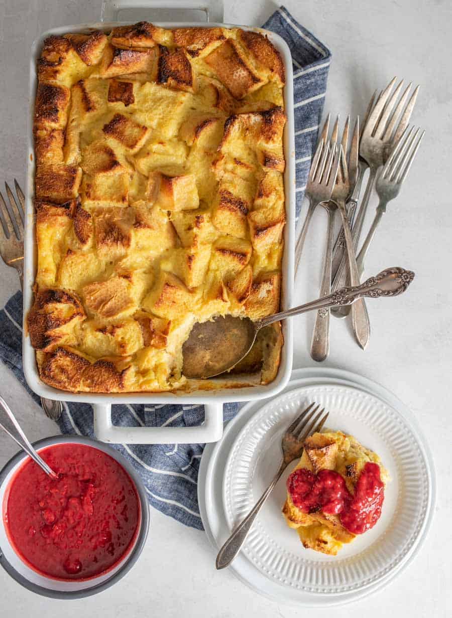 Top view of French toast breakfast casserole with a portion of it on a plate with raspberry sauce on top and a bowl of raspberry sauce next to it. 
