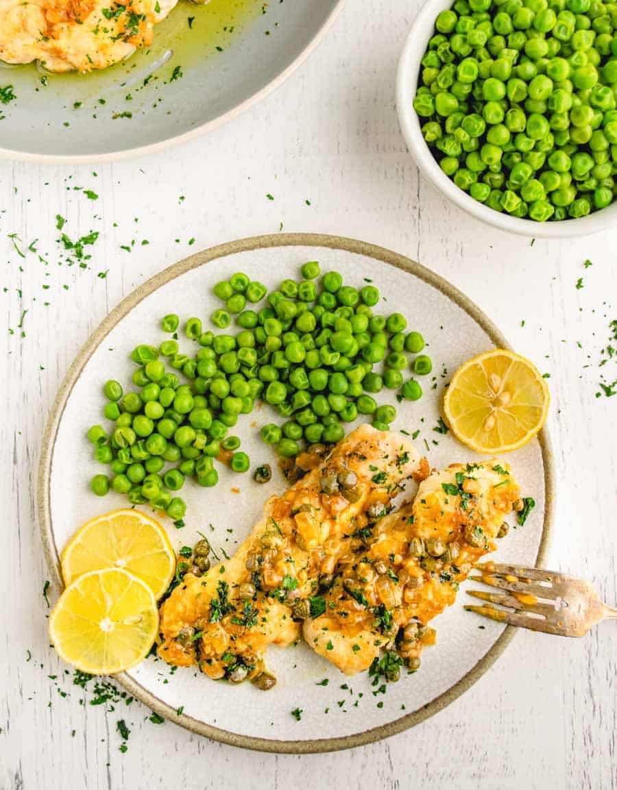 A plate of chicken picatta with lemon slices and a side of peas. 
