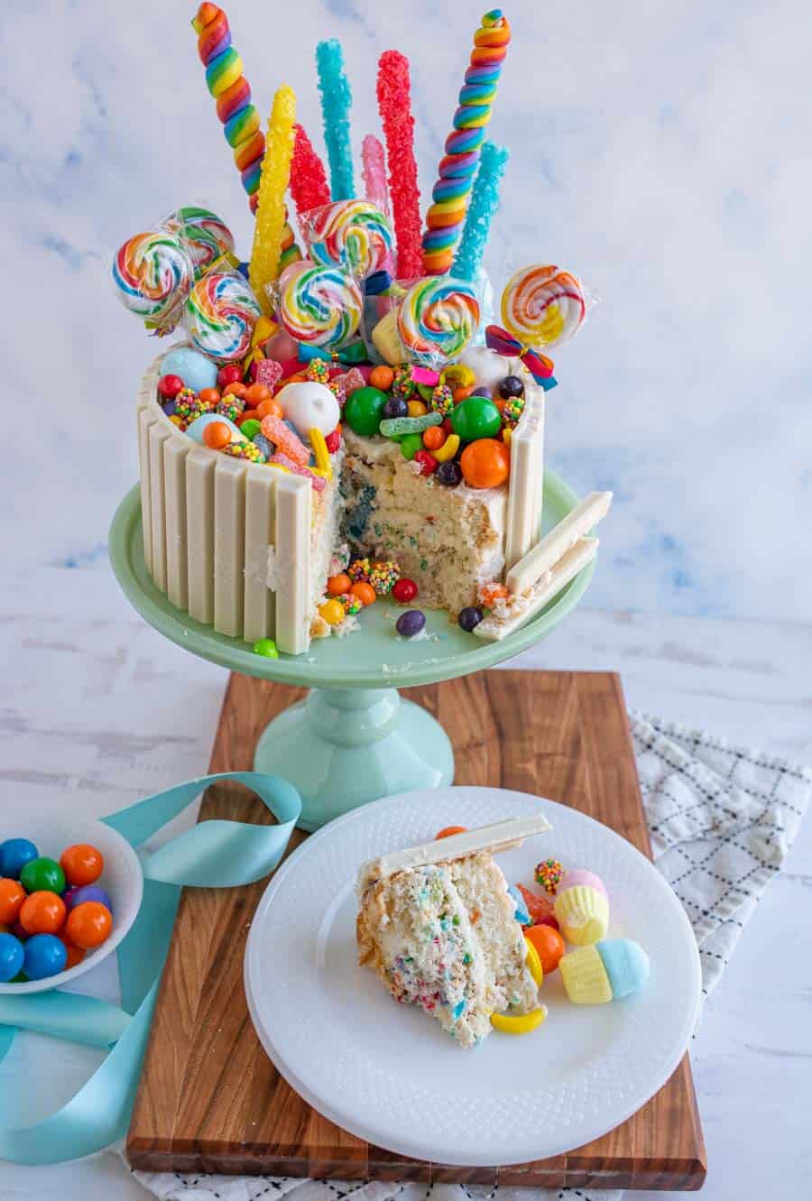 picture of a slice of candy cake on a plate with the cake on a cake stand in the background