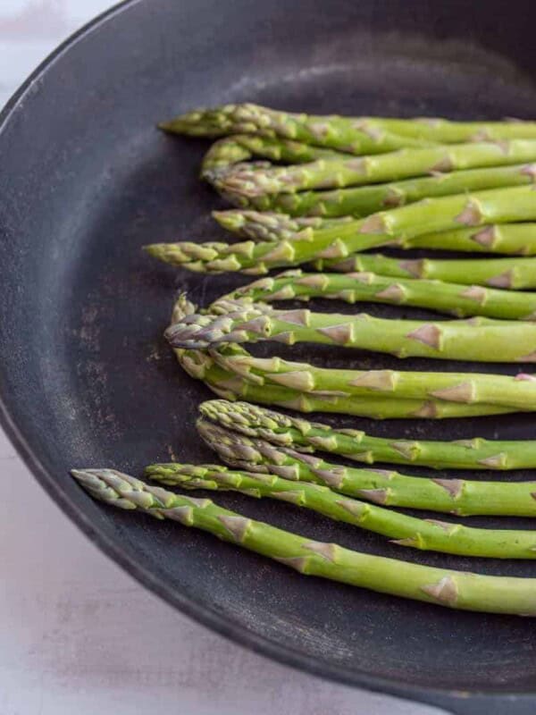 A bunch of fresh green asparagus spears, perfectly sautéed, arranged in a black cast iron pan on a light, textured surface.