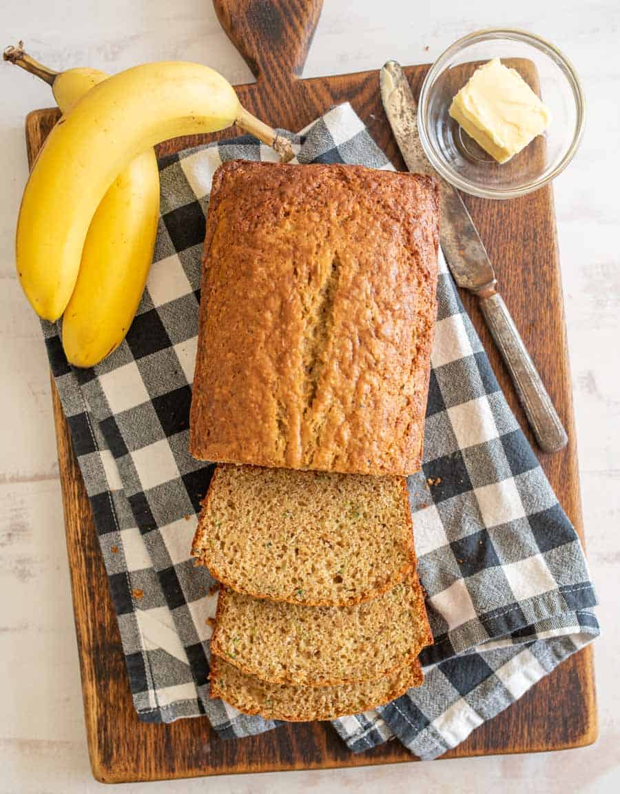 Top view of banana bread with slices cut from it next to bananas and a bowl of butter