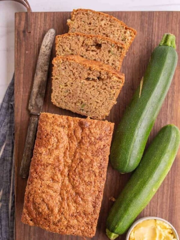 top view of a loaf of zucchini bread with some slices cut out of it next to zucchinis and a bowl of butter