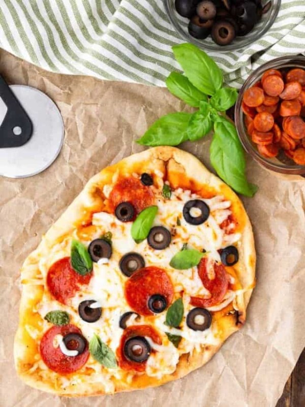 top view of flatbread pizza next to a bowl of pepperonis, olives, and basil leaves