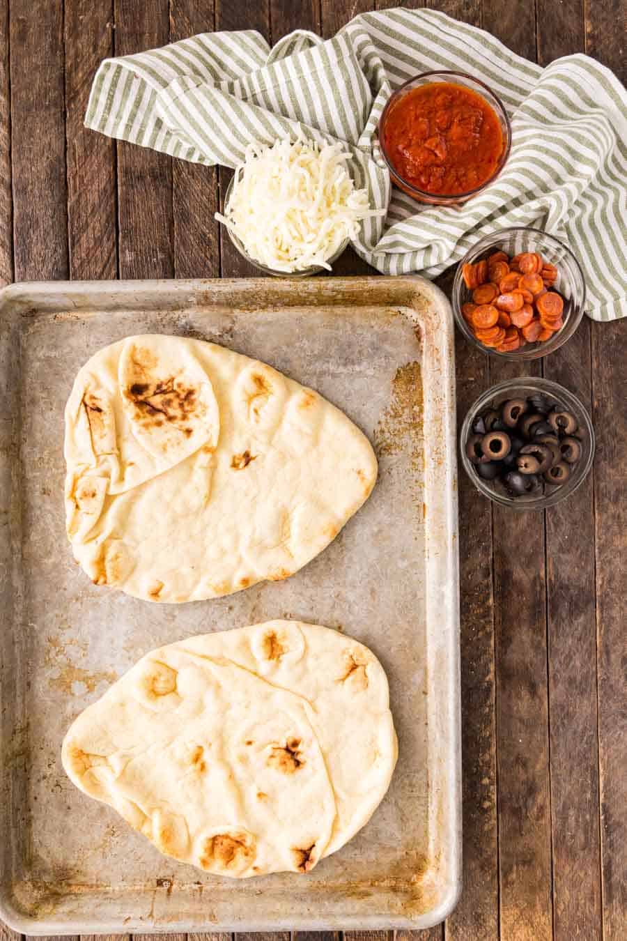 top view of flatbread on a baking sheet next to bowls of toppings for pizza.