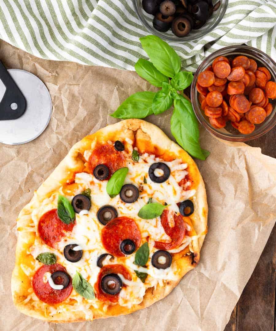 top view of flatbread pizza next to a bowl of pepperonis, olives, and basil leaves.