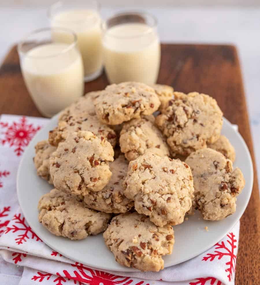 A Christmas setting on wooden block with egg nog and traditional tan looking pecan sandies cookies on a white plate. 