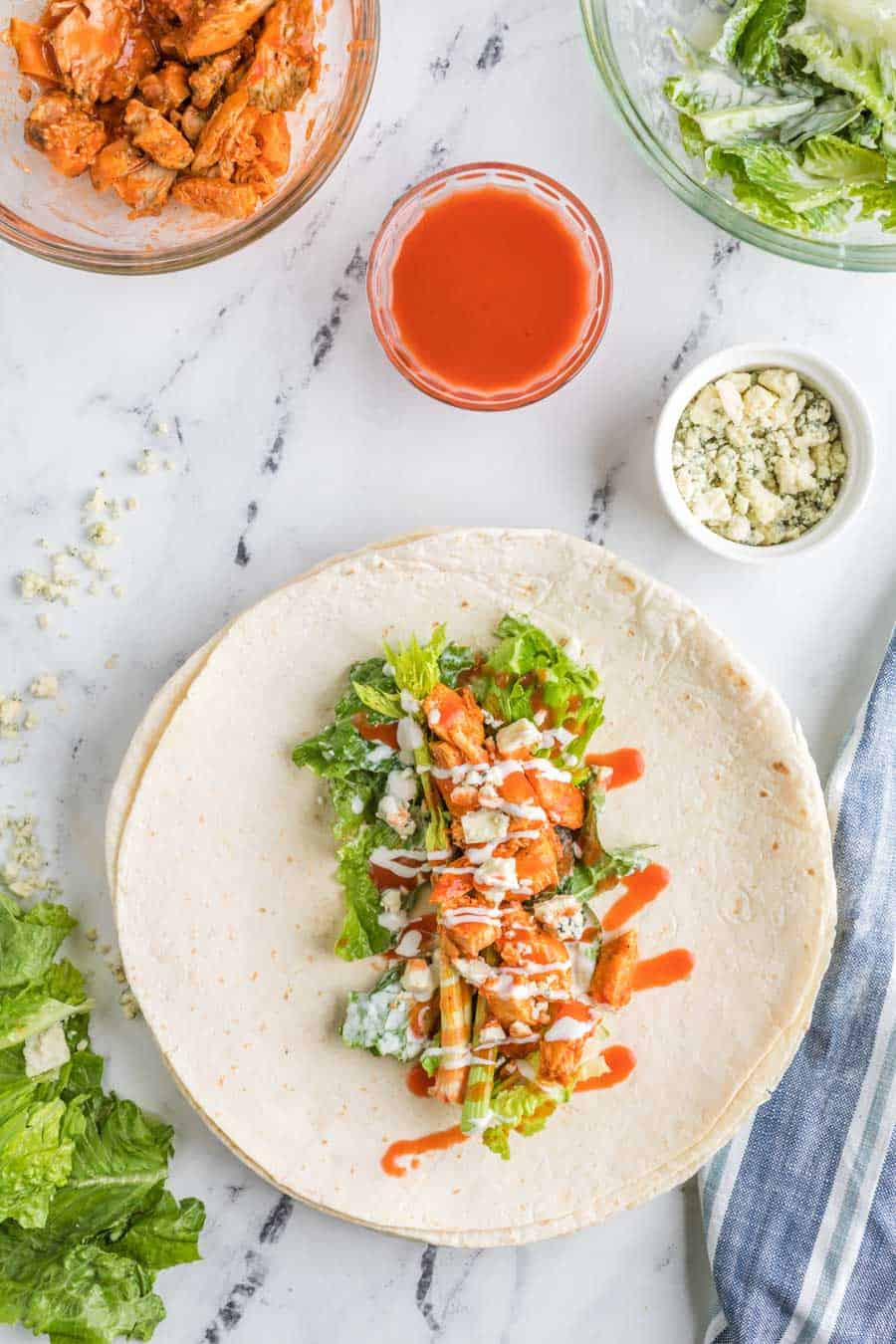 Top view open flour tortilla with buffalo chicken wrap ingredients on the inside with a squirt of dressing of it and all ingredients also in bowls arranged around. 