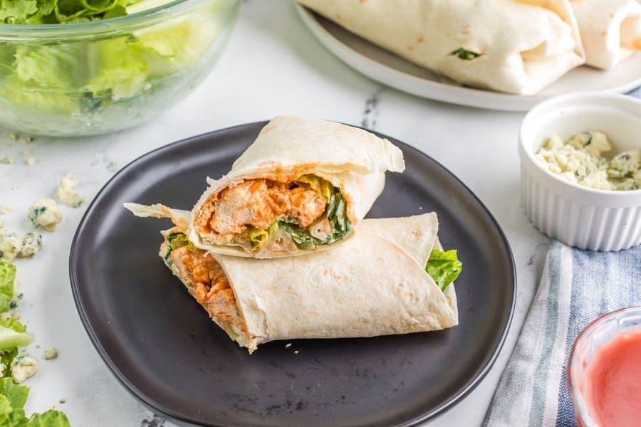 A black plate, buffalo chicken wraps - flour tortilla wrapped saucy cooked chicken and lettuce sliced to see what's inside. 