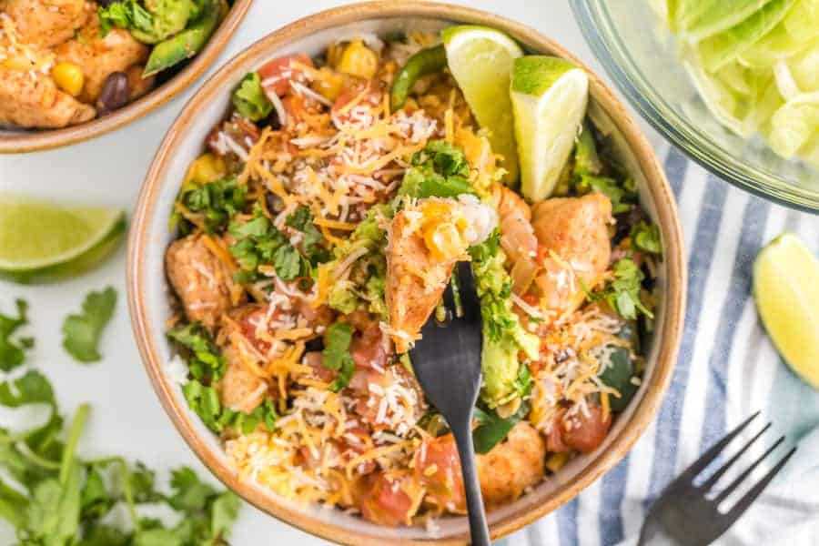 A very close view of chicken burrito bowl to see detail of limes and cilantro and cheese avocados and tomatoes with fork. 