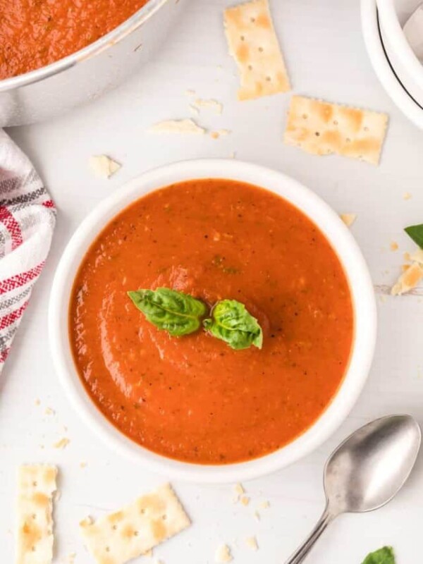 top view of roasted tomato soup in a white cup with crackers, spoon, and a sprig of basil