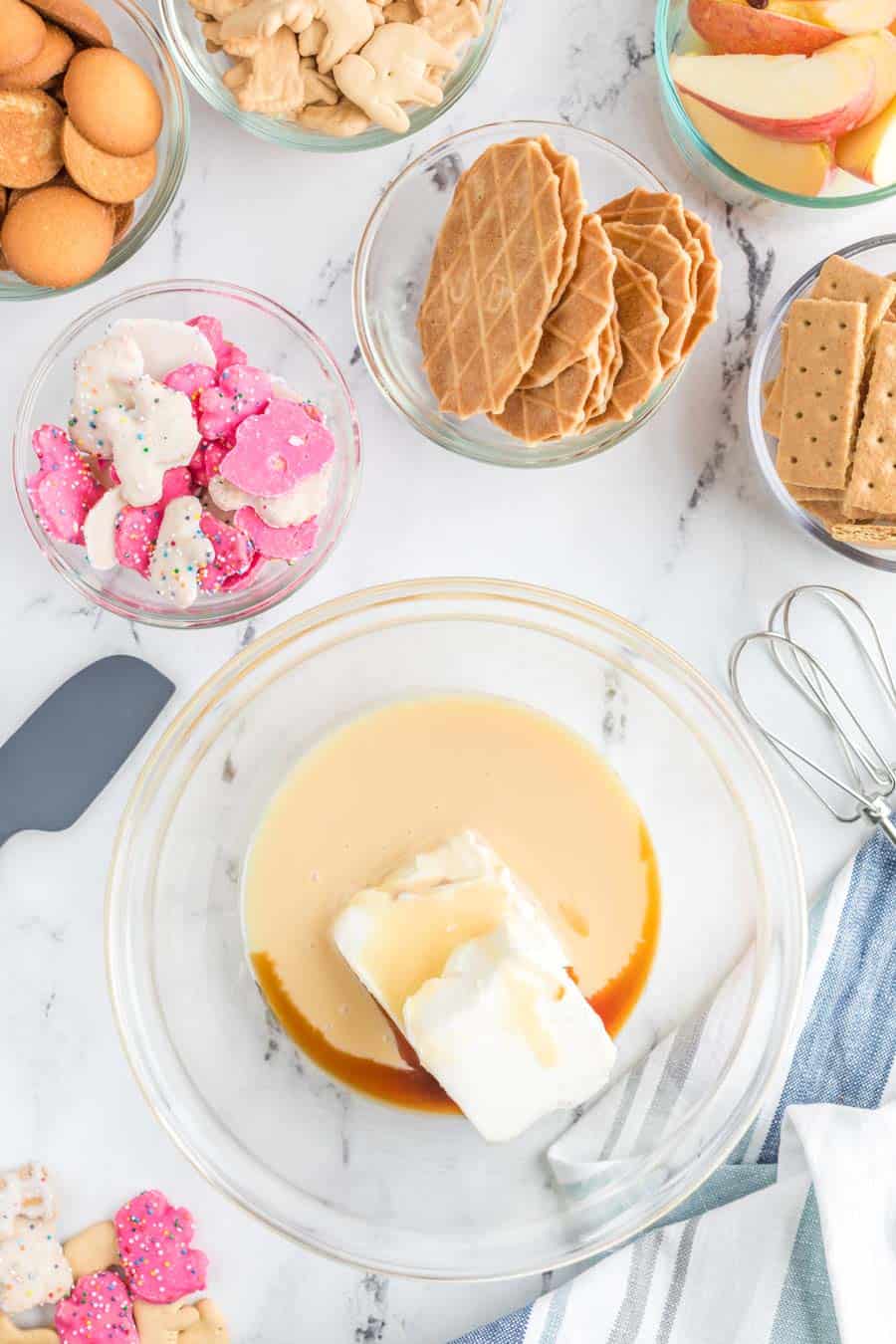 top view of snacks and other dip-able foods such as gram crackers, apples, and wafers surrounding a glass mixing bowl with cream and vanilla and a block of cream cheese