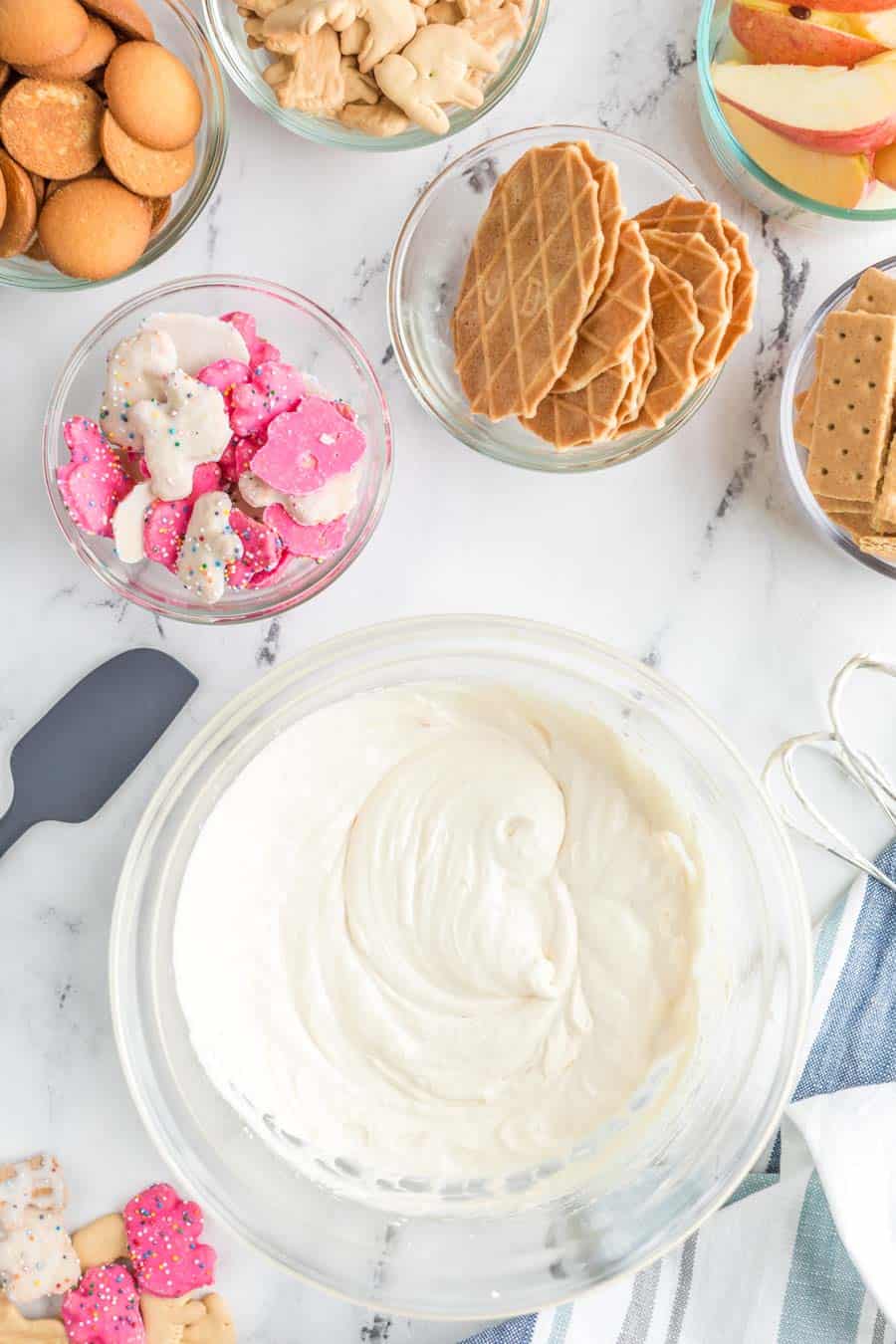 top view of snacks and other dip-able foods such as gram crackers, apples, and wafers surrounding a glass bowl with a silky white cheesecake dip in a mixing bowl