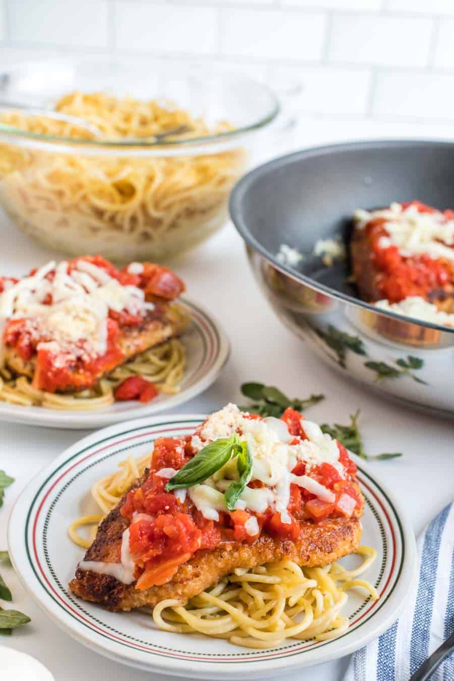 Whole scene with multiple plates and a bowl of cooked pasta and a pan - top view chicken parmesan dinner plate - spaghetti pasta and breaded chicken breast with red sauce on top on a white and Italian themed table