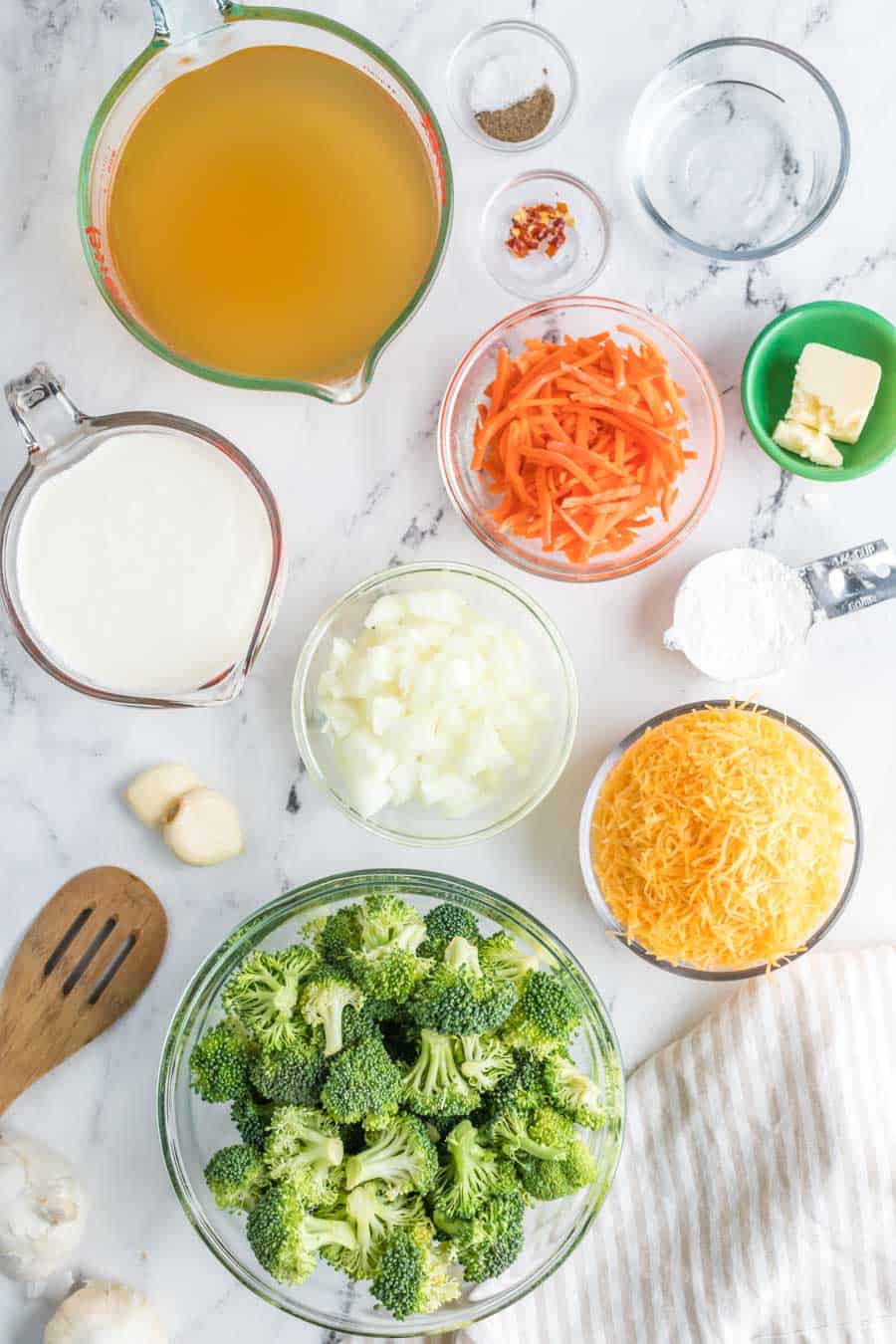 Top view of raw ingredients of the broccoli cheddar soup in pyrax and glass bowls over a marble counter. 