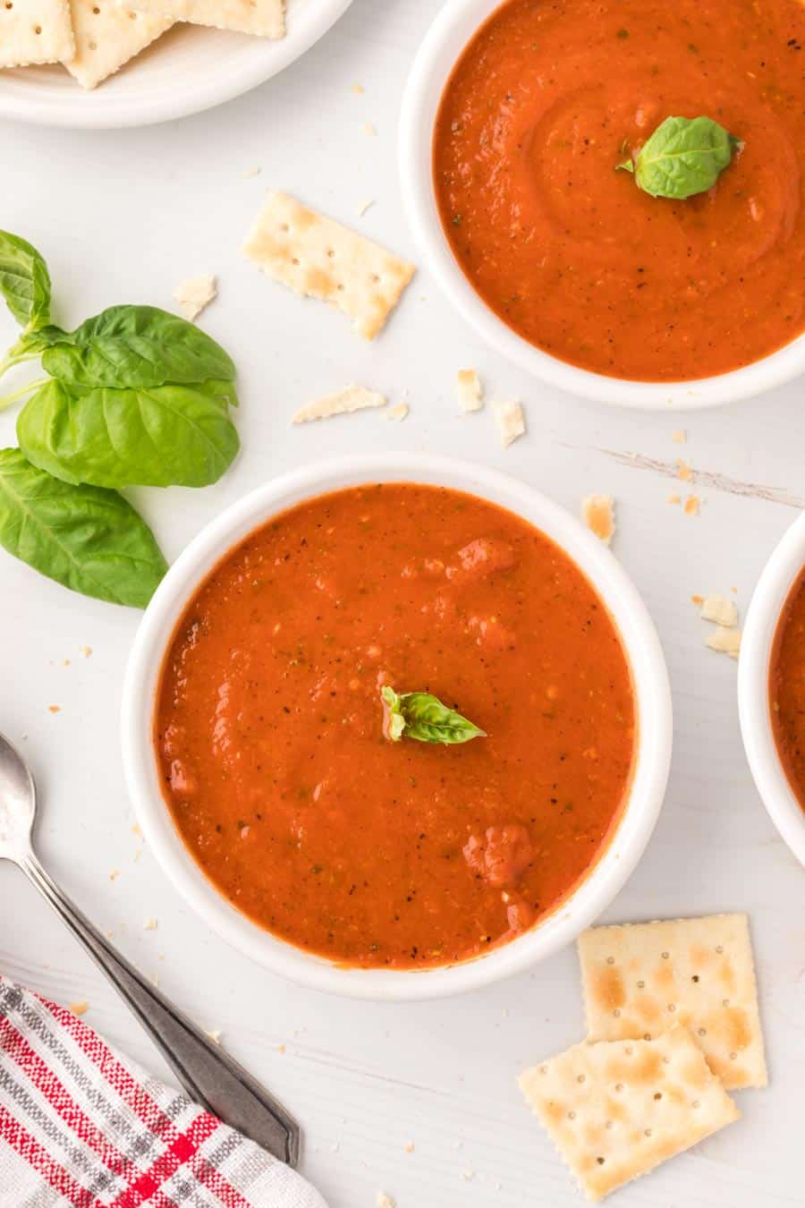 Top view of roasted tomato soup in a white cup with crackers, spoon, and a sprig of basil. 