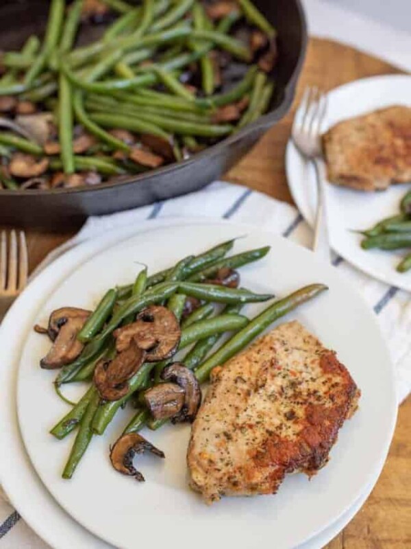 cast iron skillet with cooked long green beans and browned sliced mushrooms in the background in the foreground is a white plate with the vegetables on the side and a cooked pork chop lightly seasoned next to it