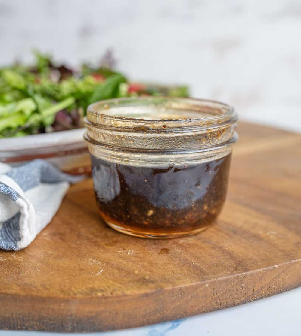 small jar with dark salad dressing in it on a brown board with salad on plate in background.