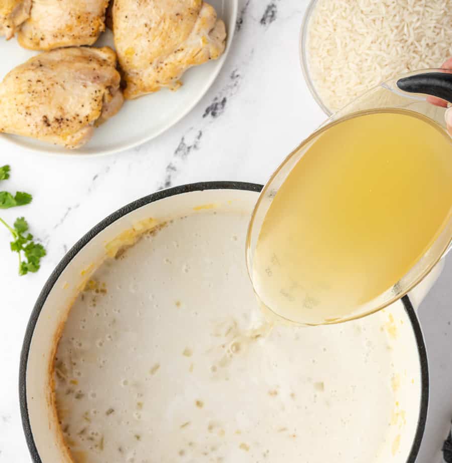 top view of cast iron pot with a coconut sauce and POV of a hand pouring broth into it with uncooked rice to the side and seasoned and cooked chicken on another plate to the side