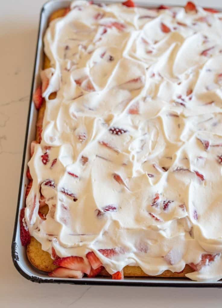 A rectangular sheet cake topped with whipped cream and slices of strawberries. The cream is spread unevenly over the cake, with visible pieces of strawberries peeking through. The cake is in a metal baking pan on a light surface.