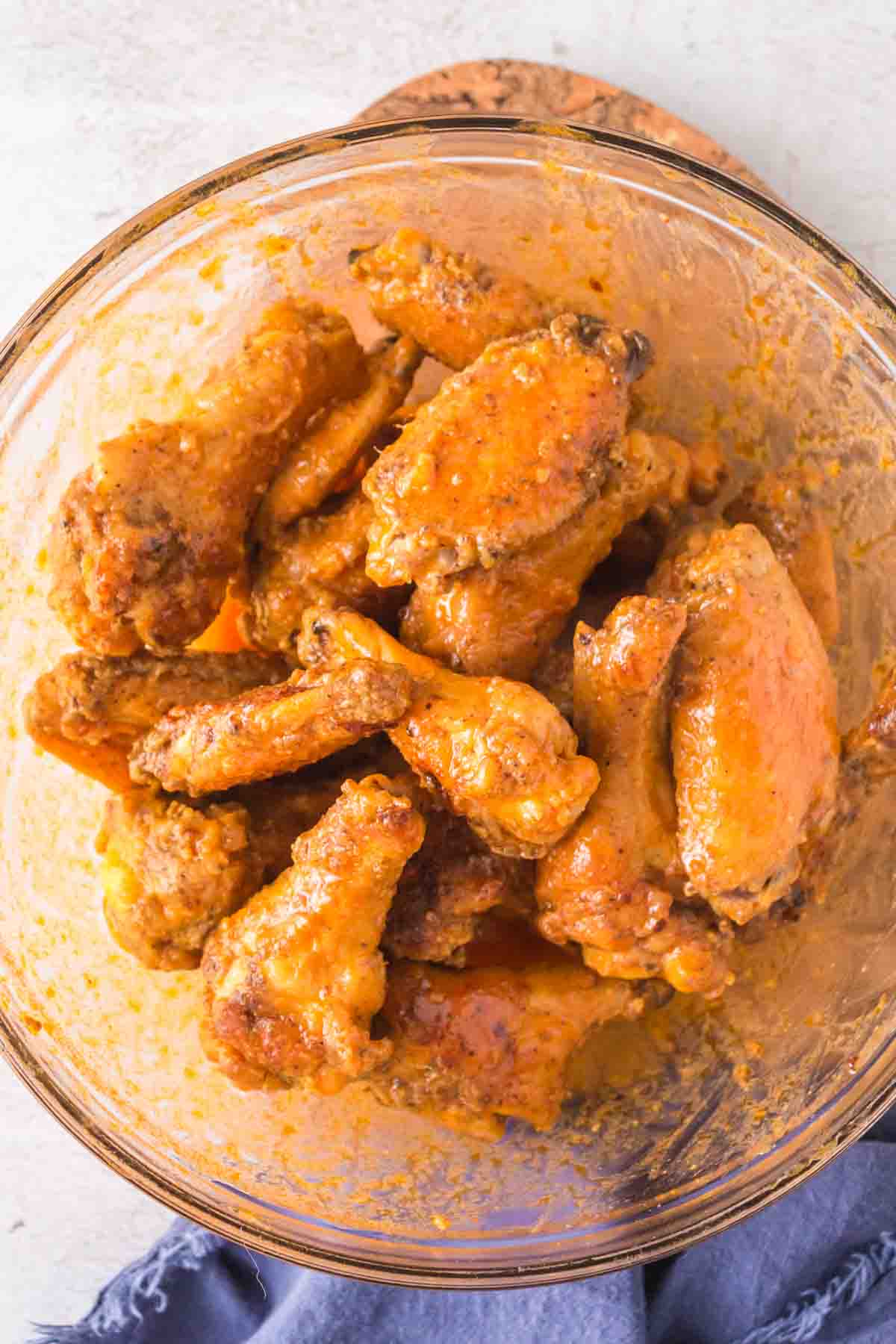 A glass bowl filled with golden-brown chicken wings coated in a thick, orange sauce. The surface beneath is light-colored, and a small piece of blue cloth is partially visible at the bottom edge.