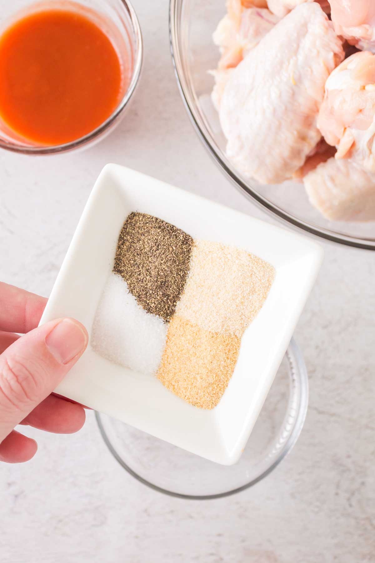 A hand holds a square dish with four spices: black pepper, salt, onion powder, and garlic powder. Nearby, a bowl contains raw chicken wings ready to transform into fiery hot wings, and another small bowl brims with orange hot sauce. The background is a light-colored surface.