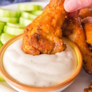 A close-up captures a hand dipping a golden-brown baked hot wings into a bowl of creamy white sauce. Sliced celery sticks sit invitingly in the background on a pristine white plate.