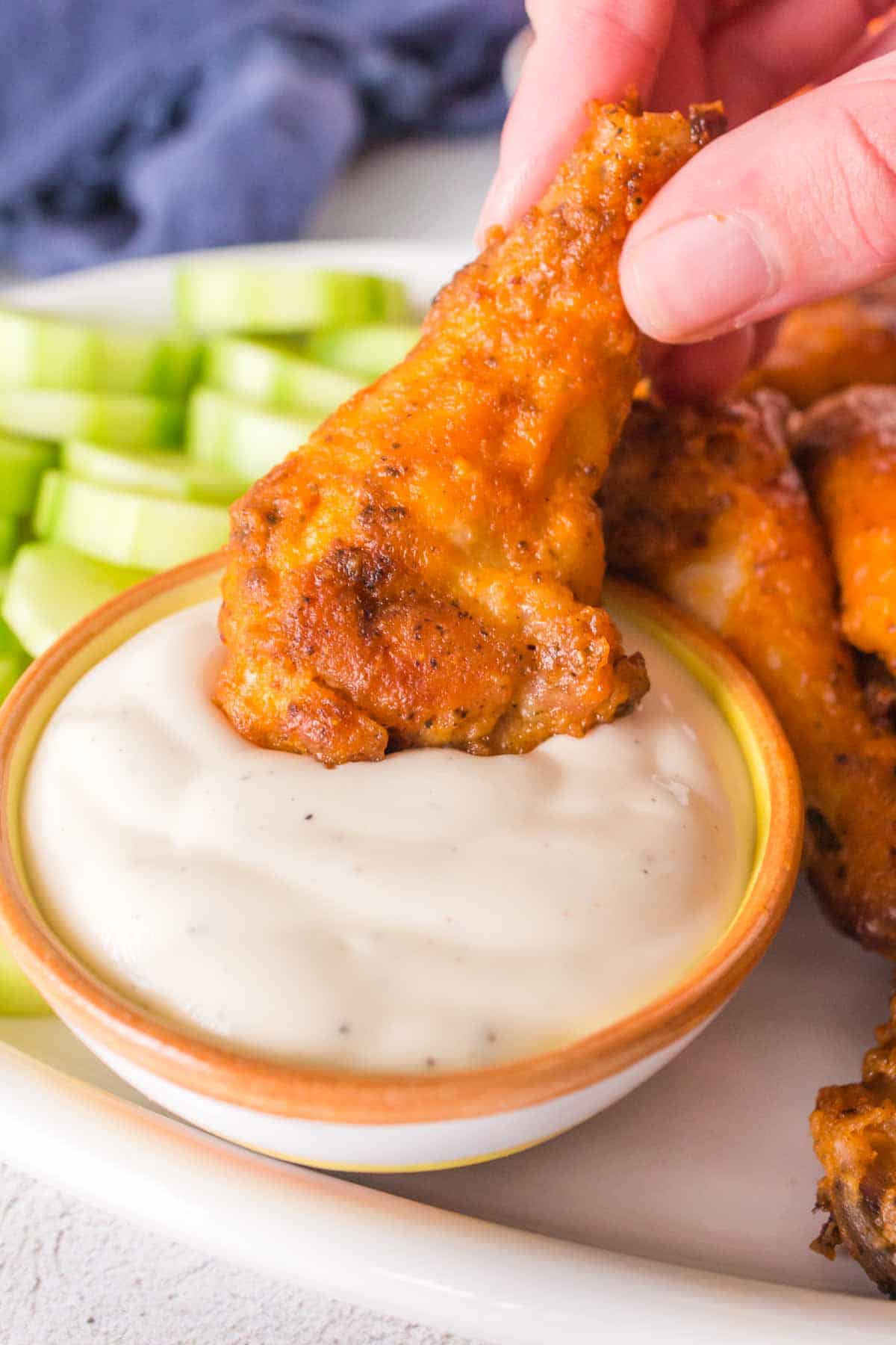 A close-up captures a hand dipping a golden-brown baked hot wings into a bowl of creamy white sauce. Sliced celery sticks sit invitingly in the background on a pristine white plate.