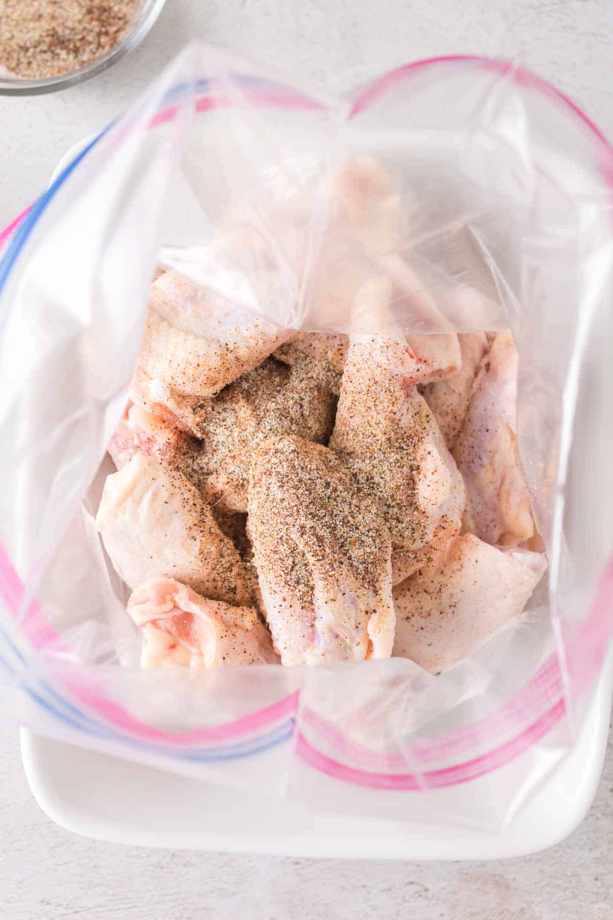 Raw chicken wings in a plastic bag with a zipper seal, sprinkled with seasoning, placed inside a white bowl on a light-colored surface.